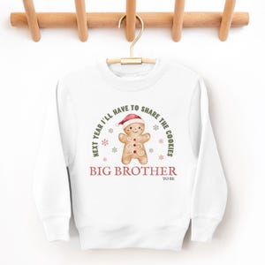 May include: White sweatshirt with a gingerbread man graphic wearing a Santa hat. The text reads "Next year I'll have to share the cookies" and "Big Brother to be". The design includes snowflakes.