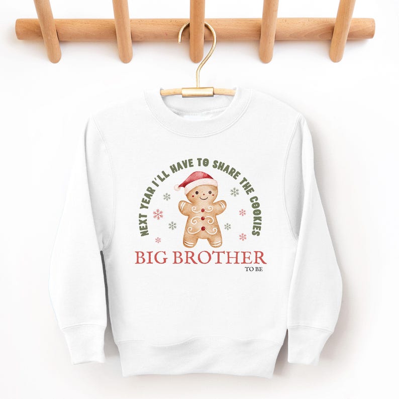 May include: White sweatshirt with a gingerbread man graphic wearing a Santa hat. The text reads "Next year I'll have to share the cookies" and "Big Brother to be". The design includes snowflakes.