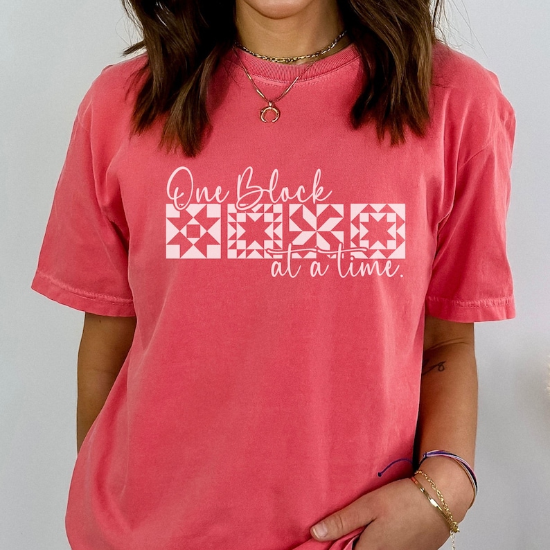 May include: Coral-colored t-shirt with the text "One Block at a Time" in white script and block letters. The design includes four white quilt block patterns. The shirt has short sleeves and a crew neck.