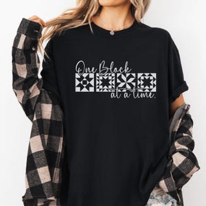 May include: Black t-shirt with the text "One Block at a Time" in white script and block designs. The shirt is paired with a black and brown plaid jacket.