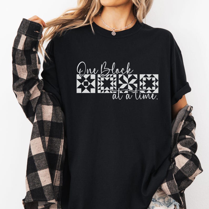 May include: Black t-shirt with the text "One Block at a Time" in white script and block designs. The shirt is paired with a black and brown plaid jacket.