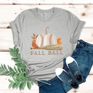 Puede incluir: Una camiseta gris con una calabaza blanca con costuras de béisbol y un bate de béisbol marrón. El texto "Fall Ball" está impreso debajo de la calabaza y el bate.