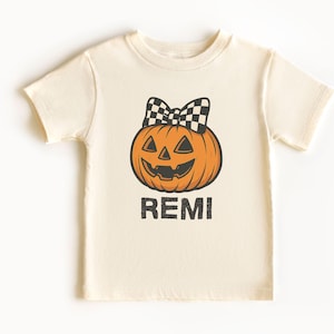 May include: A cream-coloured t-shirt with an orange pumpkin design, a black and white checkered bow, and the name "REMI" in black. This Halloween-themed t-shirt is suitable for children. The pumpkin has a smiling face.