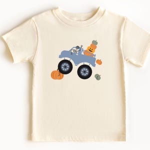 May include: Cream-coloured t-shirt with a blue monster truck driven by a skeleton, carrying pumpkins. The truck has large black wheels. Orange and green pumpkins are scattered around the truck, creating a Halloween theme.