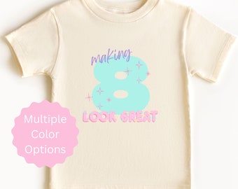 May include: A light beige t-shirt with a light blue number 8 and the text "making 8 look great" in pink and purple. The number 8 has pink and white sprinkles around it.