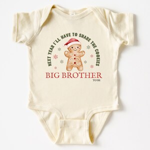 May include: Cream-colored baby bodysuit with a gingerbread man graphic wearing a Santa hat. Text reads "Next year I'll have to share the cookies" and "Big Brother to be". Festive holiday design.