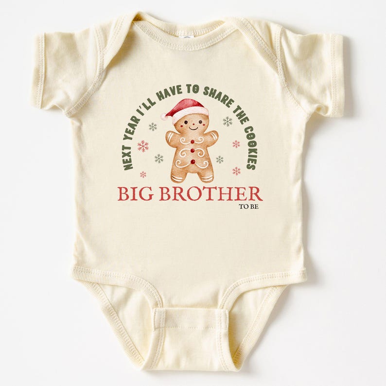 May include: Cream-colored baby bodysuit with a gingerbread man graphic wearing a Santa hat. Text reads "Next year I'll have to share the cookies" and "Big Brother to be". Festive holiday design.