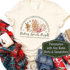 May include: A beige t-shirt with a green circle design featuring a gingerbread house, gingerbread man, and snowflakes. The text reads "Grandma's Christmas Cookie Co. Baking Spirits Bright Free Hot Cocoa Open All Season Discounts for Santa".