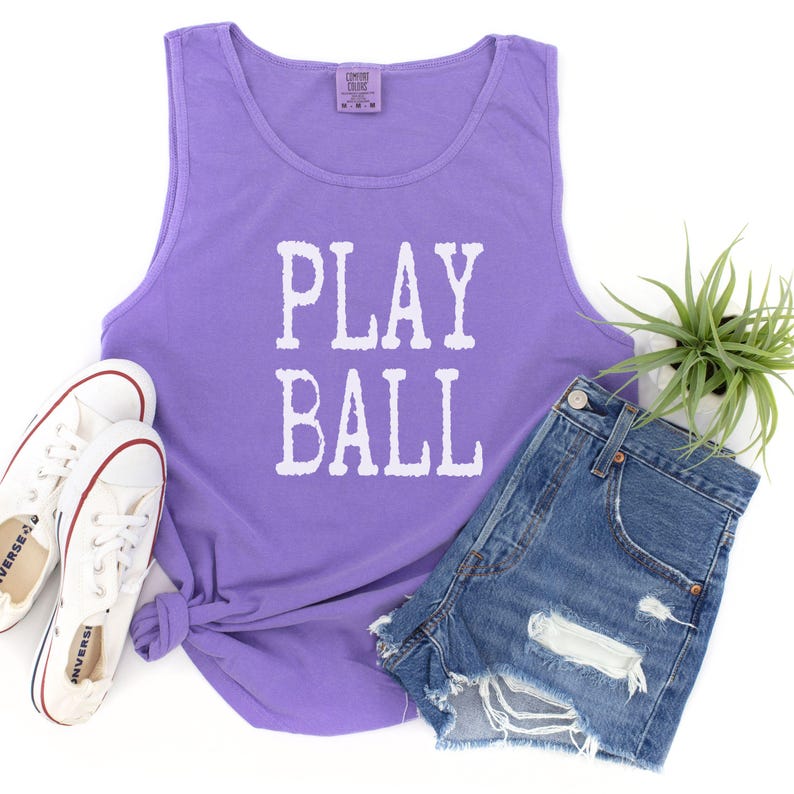 Puede incluir: Una camiseta de tirantes morada sin mangas con el texto blanco PLAY BALL impreso en la parte delantera. La camiseta se muestra junto a un par de zapatillas de lona blancas, pantalones cortos de mezclilla azul con detalles desgastados y una peque&ntilde;a planta verde en maceta.