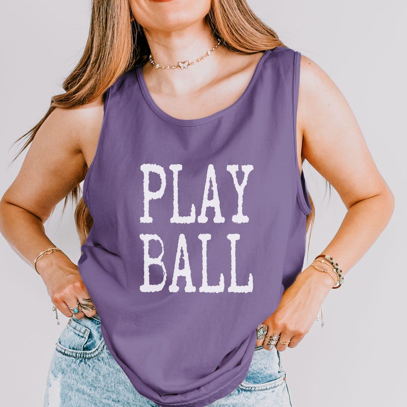 Puede incluir: Una camiseta sin mangas de color morado con el texto PLAY BALL impreso en letras blancas de bloque en el pecho. La prenda tiene un cuello redondo y se combina con unos vaqueros de mezclilla azul claro, creando un estilo casual y deportivo.