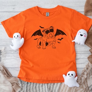 May include: Orange t-shirt with a black silhouette of a bulldog wearing sunglasses and bat wings. The bulldog is surrounded by bats. The shirt is on a wooden surface with white ghost toys.