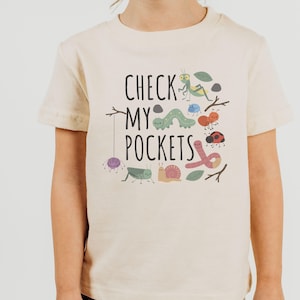 May include: Beige short-sleeved t-shirt with the text "CHECK MY POCKETS" and colorful illustrations of insects. The design includes a spider, caterpillar, ladybug, snail, and other bugs.