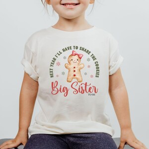 May include: A cream-colored t-shirt featuring a gingerbread person with a pink bow and the words "Next year I'll have to share the cookies" and "Big Sister to be" in red script.