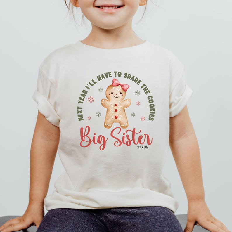 May include: A cream-colored t-shirt featuring a gingerbread person with a pink bow and the words "Next year I'll have to share the cookies" and "Big Sister to be" in red script.