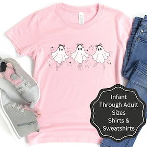 May include: A light pink t-shirt featuring a graphic of three dancing ghost figures in ballet shoes. The text on the image reads "Infant Through Adult Sizes Shirts & Sweatshirts". A pair of silver shoes and blue jeans are also in the image.