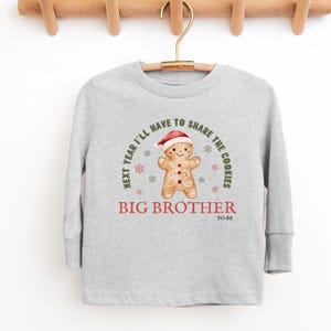 May include: A light gray long-sleeved shirt with a gingerbread man graphic wearing a Santa hat. The text reads "Next year I'll have to share the cookies" and "Big Brother to be."