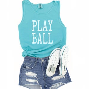 Puede incluir: Una camiseta sin mangas de color turquesa con el texto blanco PLAY BALL impreso en la parte delantera, combinada con pantalones cortos de mezclilla azul desgastados y un par de zapatillas de lona blancas con cordones y una raya azul.