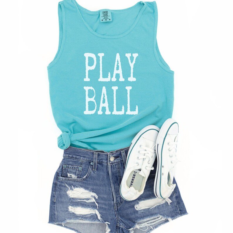 Puede incluir: Una camiseta sin mangas de color turquesa con el texto blanco PLAY BALL impreso en la parte delantera, combinada con pantalones cortos de mezclilla azul desgastados y un par de zapatillas de lona blancas con cordones y una raya azul.