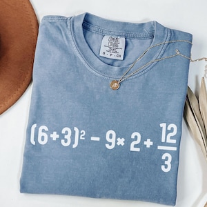 May include: A blue Comfort Colors t-shirt with white text displaying a mathematical equation. A gold necklace with a pendant rests on the shirt. A brown hat is visible in the upper left corner.