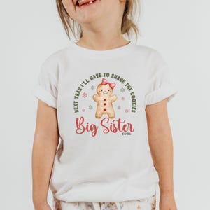 May include: White t-shirt featuring a gingerbread person with a pink bow and the text "Big Sister to be". The shirt also has the text "Next year I'll have to share the cookies" in a circular design.