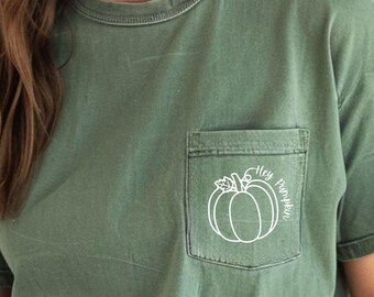 Może przedstawiać: Szałwiowo-zielony t-shirt z krótkim rękawem, okrągłym dekoltem i kieszenią na piersi. Na kieszeni znajduje się biały rysunek konturowy dyni z liściem i pnączem oraz biały napis kursywą o treści Hey Pumpkin.