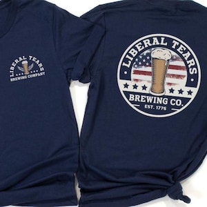 May include: Two navy blue t-shirts. One shirt features a logo with the text "LIBERAL TEARS BREWING COMPANY" and a beer glass with wings. The other shirt has a circular design with a beer glass over the American flag and the text "LIBERAL TEARS BREWING CO. EST. 1776."