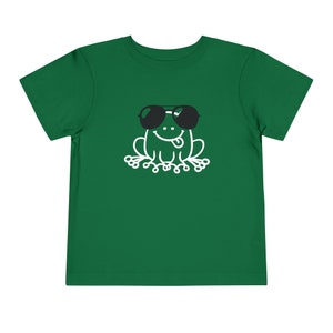 May include: A green t-shirt with a white cartoon frog wearing sunglasses. The frog is sitting with its tongue sticking out.