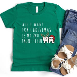 May include: Green t-shirt with the text "ALL I WANT FOR CHRISTMAS IS MY TWO FRONT TEETH". The graphic features two cartoon teeth, one with reindeer antlers and the other wearing a Santa hat. The shirt is paired with blue jeans and white sandals.