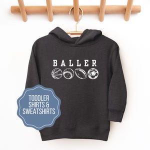 May include: A dark gray hooded sweatshirt with the word "BALLER" printed in white on the front.  Five white illustrations of different sports balls are printed below the word.  The text "TODDLER SHIRTS & SWEATSHIRTS" is printed in white on a blue circle in the lower left corner of the image.