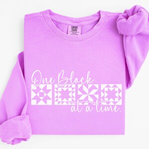May include: A light purple sweatshirt with the text "One Block at a Time" in white script. Below the text are four white quilt block designs. The sweatshirt has a crew neck and long sleeves.