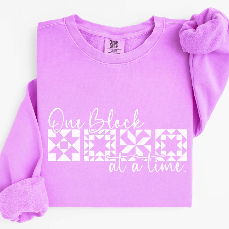 May include: A light purple sweatshirt with the text "One Block at a Time" in white script. Below the text are four white quilt block designs. The sweatshirt has a crew neck and long sleeves.