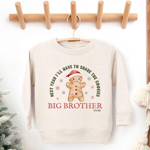 May include: A beige sweatshirt with a gingerbread man graphic wearing a Santa hat. The text reads "Next year I'll have to share the cookies" and "Big Brother to be". The sweatshirt is hanging on a wooden hanger.
