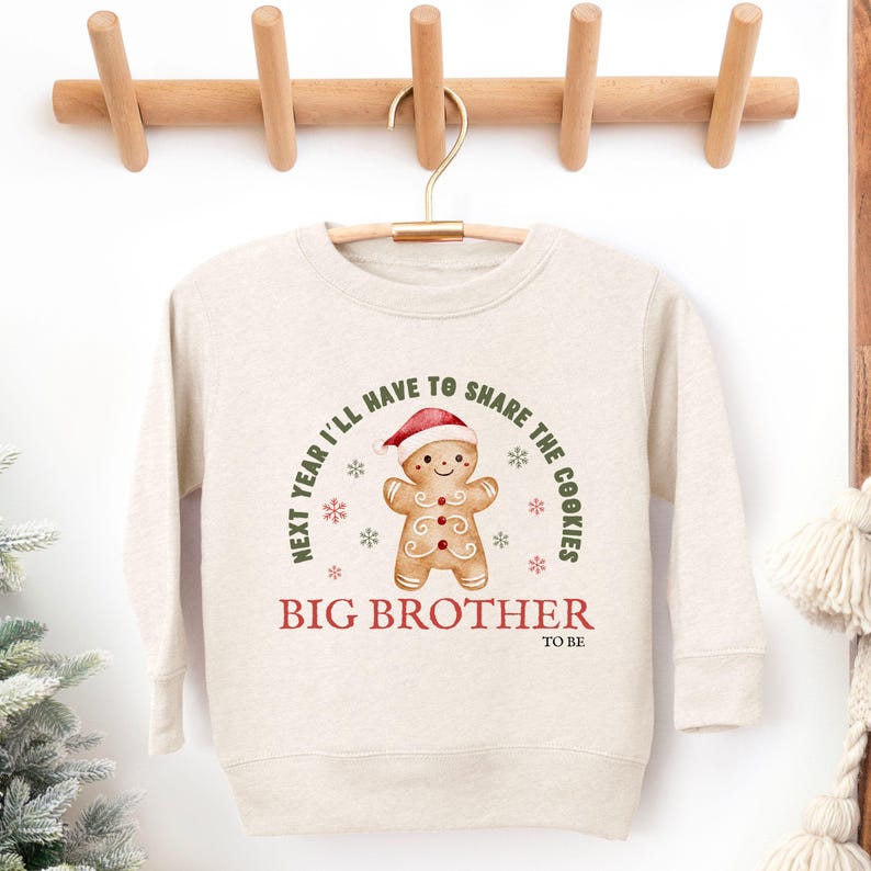 May include: A beige sweatshirt with a gingerbread man graphic wearing a Santa hat. The text reads "Next year I'll have to share the cookies" and "Big Brother to be". The sweatshirt is hanging on a wooden hanger.