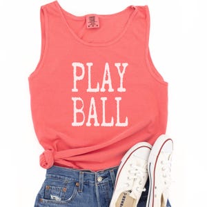 Puede incluir: Una camiseta sin mangas de color coral con el texto blanco PLAY BALL impreso en la parte delantera. La prenda se combina con pantalones cortos de mezclilla azul y zapatillas de lona blancas con ribetes rojos y azules para un estilo casual.