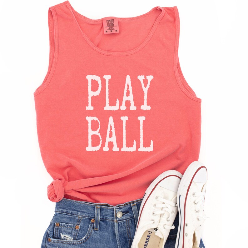 Puede incluir: Una camiseta sin mangas de color coral con el texto blanco PLAY BALL impreso en la parte delantera. La prenda se combina con pantalones cortos de mezclilla azul y zapatillas de lona blancas con ribetes rojos y azules para un estilo casual.