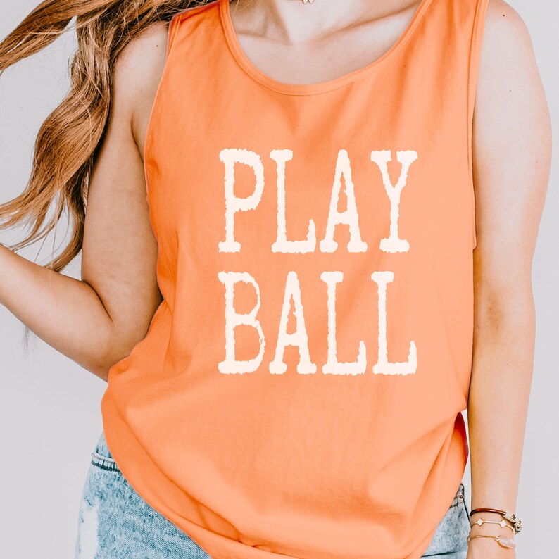 Puede incluir: Una camiseta de tirantes naranja sin mangas con el texto PLAY BALL impreso en una fuente blanca de estilo bloque desgastado en el centro del pecho. La prenda tiene un cuello redondo y un corte relajado, confeccionada en tela lisa.