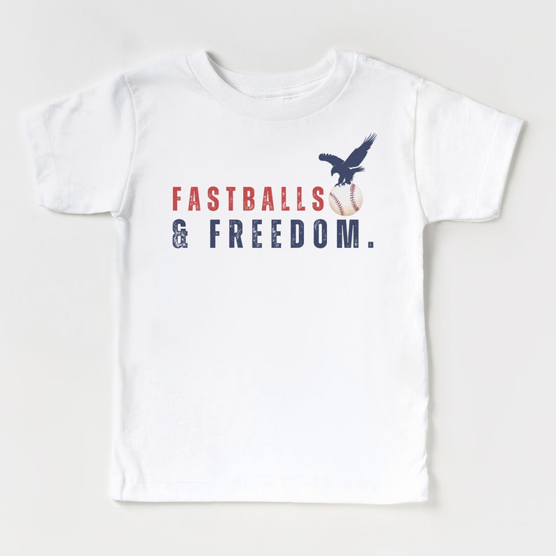 Może przedstawiać: Biała koszulka z niebieskim orłem trzymającym piłkę baseballową i tekstem "FASTBALLS & FREEDOM."