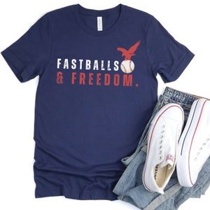 Może przedstawiać: Koszulka w kolorze granatowym z napisem "Fastballs & Freedom." w kolorze czerwonym i białym. Piłka baseballowa w kolorze czerwonym, białym i niebieskim z orłem siedzącym na g&oacute;rze znajduje się nad napisem.