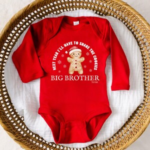 May include: A red long-sleeve baby bodysuit with a gingerbread man graphic wearing a Santa hat. The text reads "Next year I'll have to share the cookies" and "Big Brother to be". The bodysuit is displayed in a woven basket.
