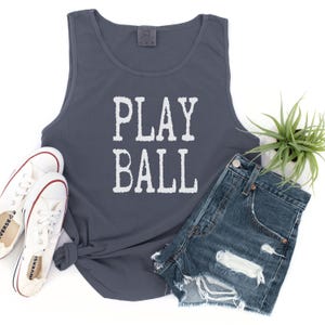 Puede incluir: Una camiseta de tirantes gris oscuro con el texto blanco PLAY BALL impreso en la parte delantera. La prenda se muestra junto a un par de zapatillas de lona blancas con ribetes rojos y azules y unos pantalones cortos de mezclilla azul desgastados.