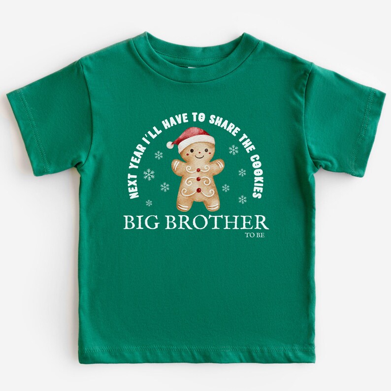 May include: A green toddler t-shirt with the text "NEXT YEAR I'LL HAVE TO SHARE THE COOKIES" and "BIG BROTHER TO BE". The shirt features a gingerbread man wearing a Santa hat and snowflakes.