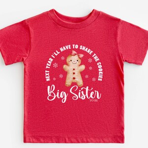 May include: A red t-shirt with a gingerbread person graphic and the text "Next year I'll have to share the cookies" and "Big Sister to be" in white. The gingerbread person has a pink bow and red buttons.