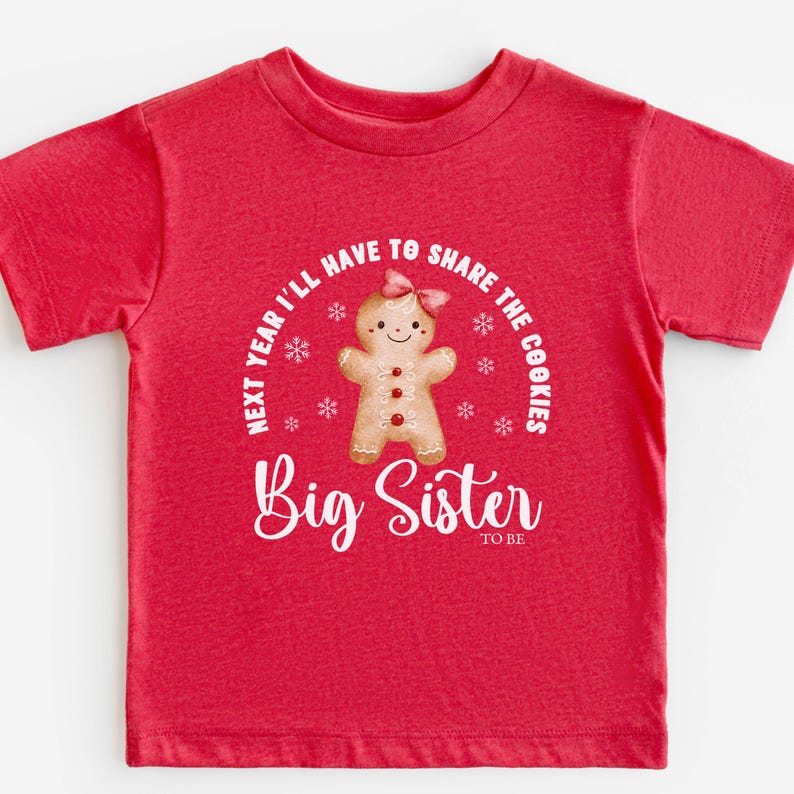 May include: A red t-shirt with a gingerbread person graphic and the text "Next year I'll have to share the cookies" and "Big Sister to be" in white. The gingerbread person has a pink bow and red buttons.
