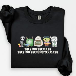 May include: Black sweatshirt featuring a line of cartoon monsters. The monsters include a skeleton, a furry green creature, a ghost with a chalkboard, a Frankenstein's monster, and a mummy. The text "THEY DID THE MATH. THEY DID THE MONSTER MATH." is printed below.