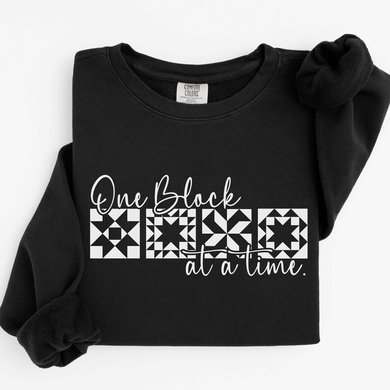 May include: Black sweatshirt with the text "One Block at a Time" in white script and block designs. The sweatshirt is made of a soft material and has a crew neck.