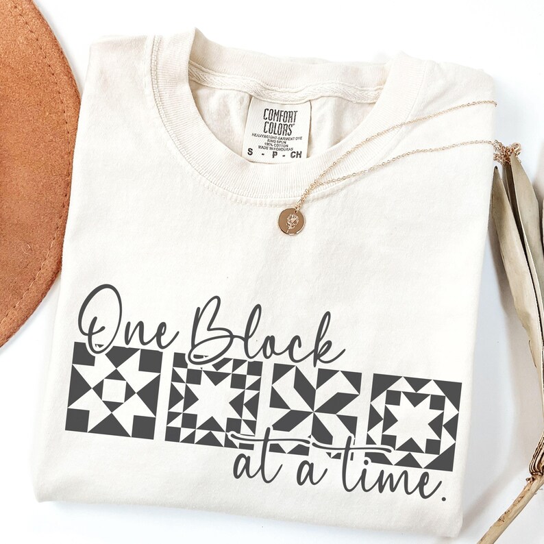May include: Cream-colored t-shirt with the text "One Block at a Time" and a geometric quilt block design in black. A gold necklace with a small pendant rests on the shirt. The shirt is a Comfort Colors brand.