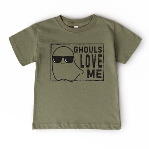 May include: Olive green t-shirt featuring a ghost wearing sunglasses with the text "GHOULS LOVE ME" in a black rectangular frame. The ghost graphic and text are in black. The shirt is a short-sleeved crew neck.