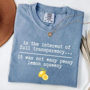 May include: A blue short-sleeved t-shirt with a white graphic print that reads, "in the interest of full transparency... it was not easy peasy lemon squeezy" above a yellow lemon illustration. The shirt features a Comfort Colors brand neck label.