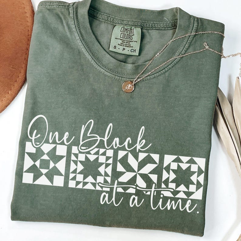 May include: Olive green t-shirt with the text "One Block at a Time" and three white quilt block designs. A gold necklace with a small pendant rests on the shirt. The shirt is folded neatly.