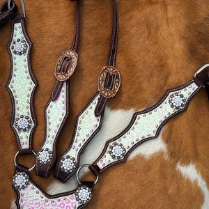 May include: A dark brown leather horse bridle and breast collar set featuring iridescent light green and pink leopard print overlays. The tack is accented with white beaded floral conchos and copper-toned oval buckles with engraved detailing.